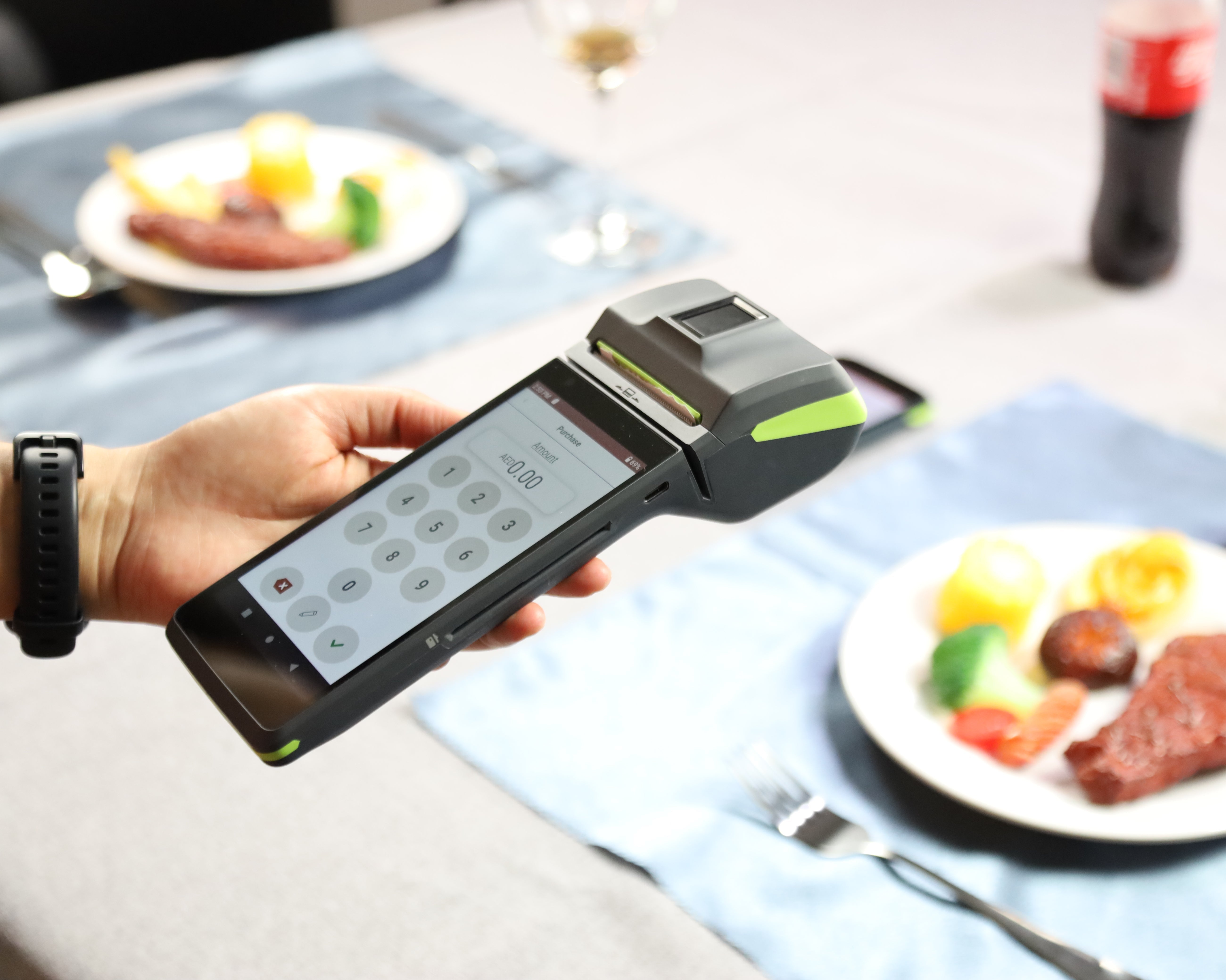 A hand holding a black and lime green handheld POS terminal with a touchscreen keypad, set against a blurred restaurant table setting with plates of food, wine glasses, and a bottle.